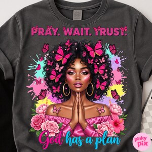 May include: Dark grey sweatshirt with a graphic of a woman adorned with butterflies. The text "PRAY. WAIT. TRUST." is above the woman, and "God has a plan" is below. The design includes pink and teal splashes and floral elements.