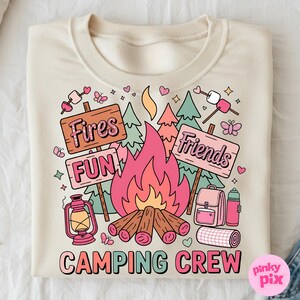 May include: Cream-colored sweatshirt with a colorful camping-themed graphic. The design includes a campfire, signs that read "Fires", "Fun", and "Friends", and the words "Camping Crew". Other elements include a lantern, backpack, and water bottle.
