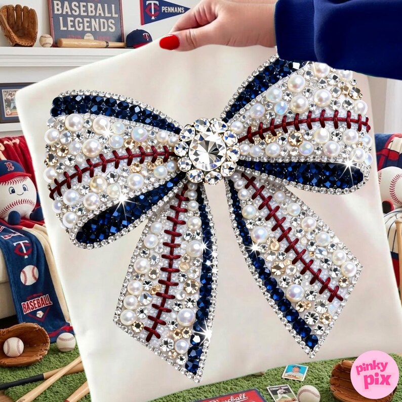 May include: A white decorative pillow featuring a large bow design. The bow is embellished with pearls, rhinestones, and dark blue accents, with red stitching details resembling a baseball. A large, sparkling crystal is at the center.
