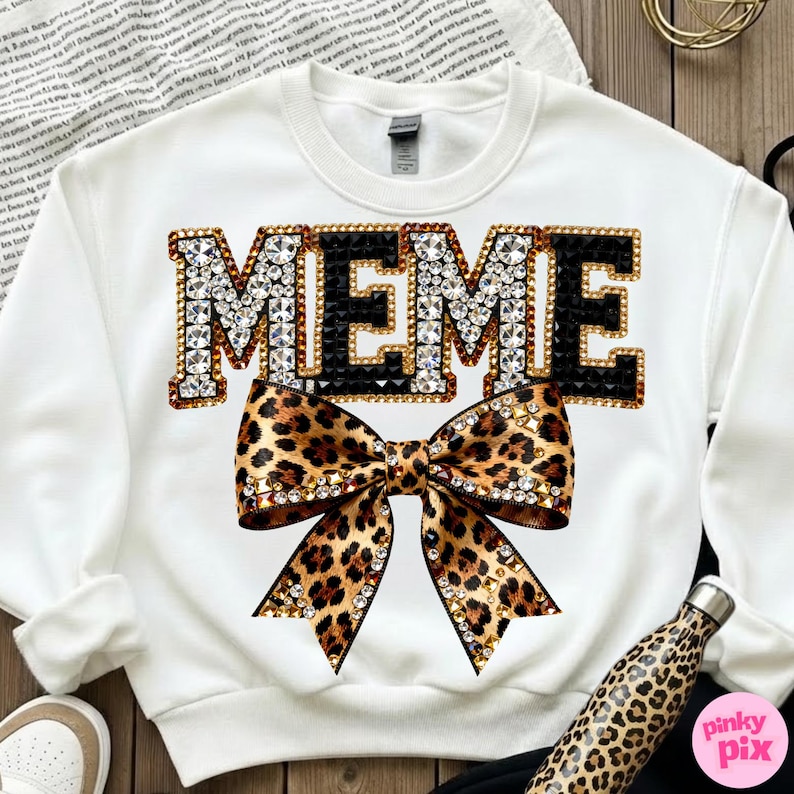 May include: White sweatshirt with the word "MEME" in large, jeweled letters. A leopard print bow is centered below the word. The sweatshirt is a casual style, suitable for everyday wear.