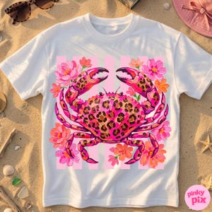 May include: White t-shirt featuring a pink crab with a leopard print design, surrounded by pink and orange flowers. The crab is set against a pink striped background. The shirt has a round neckline and short sleeves.