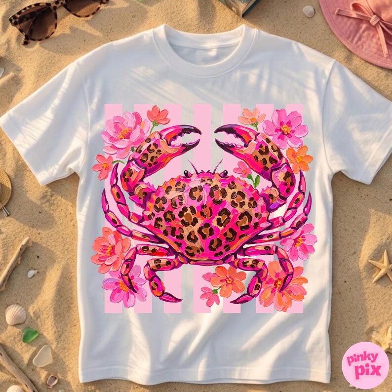 May include: White t-shirt featuring a pink crab with a leopard print design, surrounded by pink and orange flowers. The crab is set against a pink striped background. The shirt has a round neckline and short sleeves.