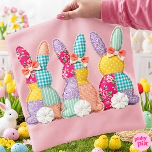 May include: A pink fabric square featuring three patchwork bunny designs. Each bunny has a different patterned ear and body, with a bow and a white flower tail. The bunnies are in shades of pink, yellow, purple, and blue.