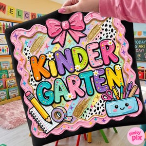 May include: A colorful "Kindergarten" sign with a pink bow, rainbow stripes, and school supplies. The sign has a black background with a pink wavy border and gold accents. The text "Kindergarten" is in large, multi-colored letters.