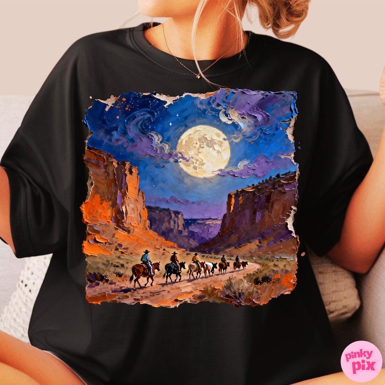 May include: Black t-shirt featuring a vibrant painting of a desert canyon scene under a full moon. Cowboys on horseback ride along a path. The artwork includes shades of blue, orange, and purple, creating a dramatic night landscape.
