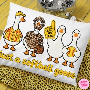 May include: White pillow with embroidered cartoon geese in baseball attire. One goose wears a baseball jersey, another a leopard print uniform, and others hold a foam finger and a softball. Text reads "just a softball goose."