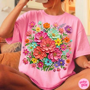 May include: A pink t-shirt featuring a vibrant floral design. The design showcases a bouquet of colorful flowers in shades of pink, orange, yellow, blue, and purple. The flowers appear to be 3D, adding depth to the design. The text "pinky pix" is visible on the bottom right.