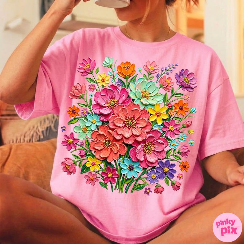 May include: A pink t-shirt featuring a vibrant floral design. The design showcases a bouquet of colorful flowers in shades of pink, orange, yellow, blue, and purple. The flowers appear to be 3D, adding depth to the design. The text "pinky pix" is visible on the bottom right.