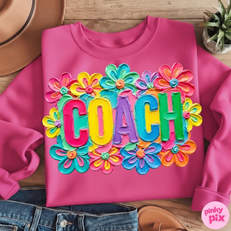 May include: A pink sweatshirt with the word "COACH" in colorful, 3D letters, surrounded by vibrant, multi-colored flowers. The sweatshirt is displayed on a wooden surface with a hat, jeans, and sandals.