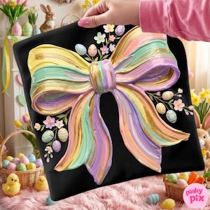 May include: Black square pillow featuring a large, textured bow in pastel colors of yellow, green, purple, and gold. The bow is adorned with painted Easter eggs and floral accents. The pillow is held by a person with a pink shirt.