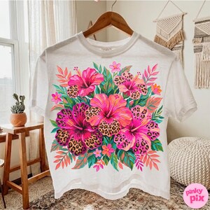 May include: White t-shirt featuring a vibrant floral design. The design showcases a bouquet of pink and orange hibiscus flowers with leopard print centers, surrounded by green leaves and pink accents. The shirt is hanging on a wooden hanger.