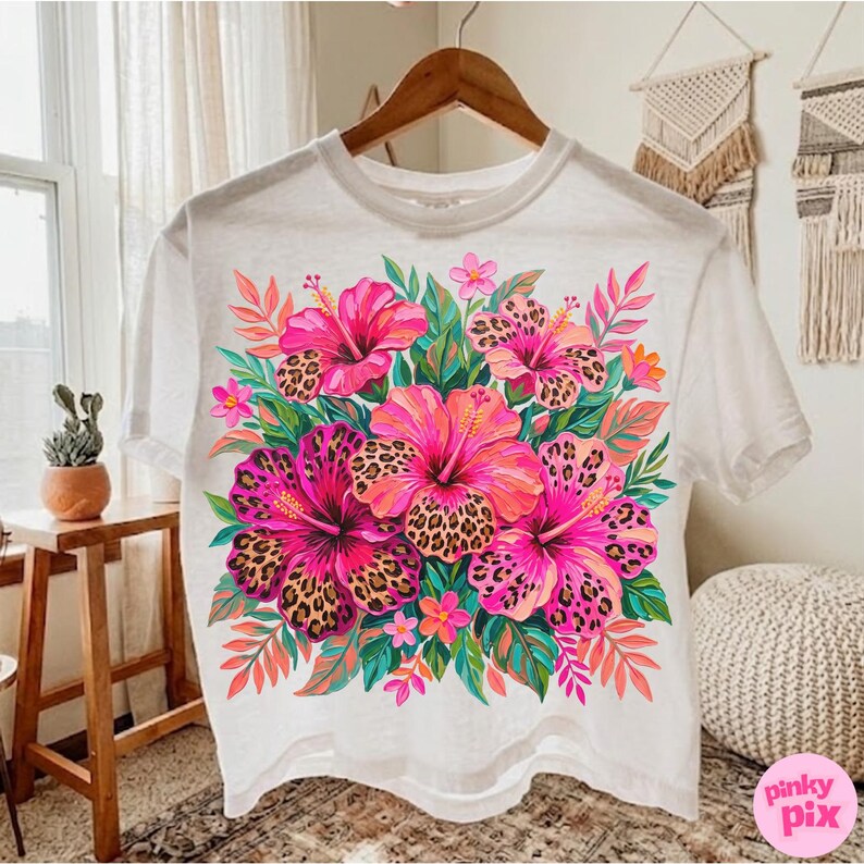 May include: White t-shirt featuring a vibrant floral design. The design showcases a bouquet of pink and orange hibiscus flowers with leopard print centers, surrounded by green leaves and pink accents. The shirt is hanging on a wooden hanger.