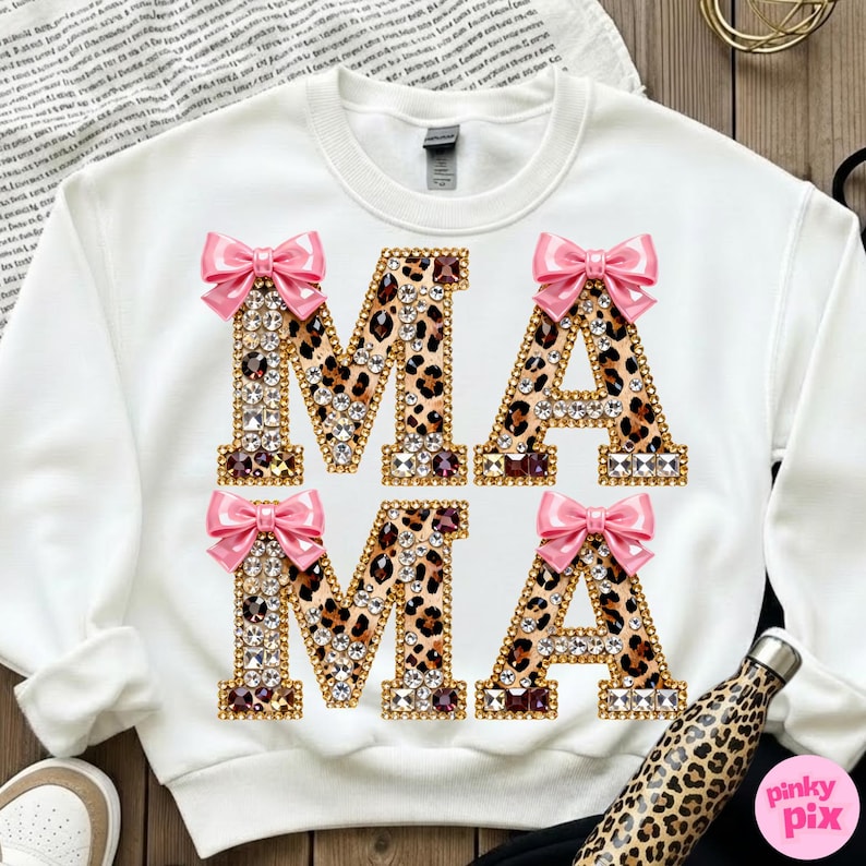 May include: White sweatshirt featuring the word "MAMA" in large, embellished letters. The letters have a leopard print pattern, are outlined in gold, and are decorated with rhinestones and pink bows. A leopard print water bottle is visible.