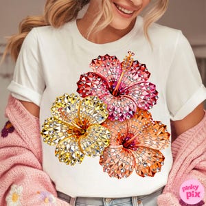 May include: White t-shirt featuring a vibrant floral design. The graphic showcases three hibiscus flowers in shades of red, orange, and yellow, each adorned with a sparkling, gem-like texture. The shirt has short sleeves and a crew neck.