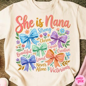 K&ouml;nnte beinhalten: Cremefarbenes T-Shirt mit dem Schriftzug "She is Nana" in Rosa und Gelb. Das Design zeigt bunte Schleifen und florale Elemente mit W&ouml;rtern wie "Strong" und "Victorious".