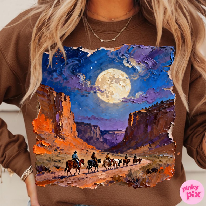 May include: Brown sweatshirt featuring a vibrant painting of a desert landscape under a full moon. The artwork depicts cowboys on horseback, riding through a canyon at night, with a palette of blues, oranges, and purples. The Pinky Pix logo is visible.