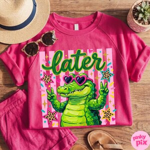 May include: A pink sweatshirt with a graphic of a green alligator wearing heart-shaped sunglasses and giving the peace sign. The word "later" is written above the alligator in green script. The design includes stars and pink stripes.