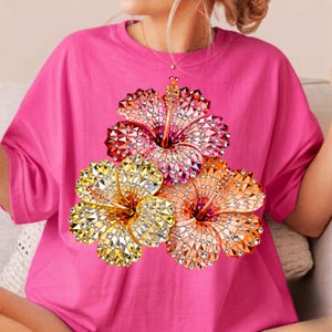 May include: A bright pink t-shirt featuring a vibrant floral design. The design showcases three hibiscus flowers in shades of red, yellow, and orange, embellished with sparkling details. The shirt has short sleeves and a relaxed fit.