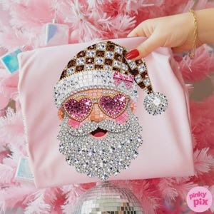 May include: A light pink fabric item featuring a decorative Santa Claus graphic composed of sparkling rhinestones. The Santa design includes a brown and white chequered hat, pink heart-shaped sunglasses, and a large, textured white crystal beard.