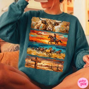 May include: Teal-colored sweatshirt featuring a multi-panel design with a bull, a cowgirl on horseback, a herd of horses, and a cross in a desert landscape. The artwork has a textured, painted appearance.