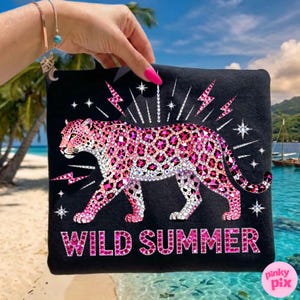 May include: Black t-shirt featuring a pink leopard design with crystal-like embellishments. The words "WILD SUMMER" are displayed in pink, with lightning bolt and star accents. The shirt is held up against a tropical beach backdrop.
