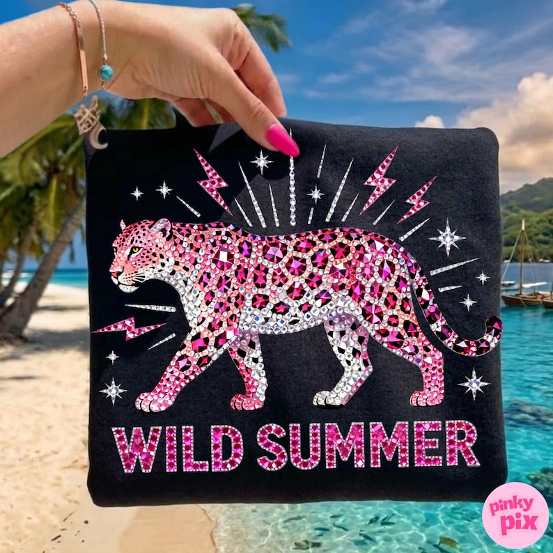 May include: Black t-shirt featuring a pink leopard design with crystal-like embellishments. The words "WILD SUMMER" are displayed in pink, with lightning bolt and star accents. The shirt is held up against a tropical beach backdrop.