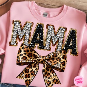 May include: Pink sweatshirt with the word "MAMA" in rhinestone-studded letters. A leopard print bow with rhinestone accents is centered below the word. The sweatshirt is a crew neck style.
