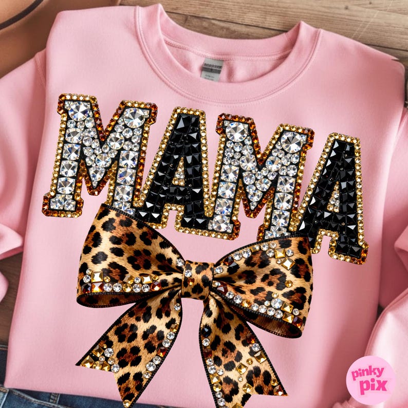 May include: Pink sweatshirt with the word "MAMA" in rhinestone-studded letters. A leopard print bow with rhinestone accents is centered below the word. The sweatshirt is a crew neck style.
