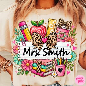 May include: A beige t-shirt featuring a colorful teacher-themed graphic. The design includes a pencil, apple, ruler, coffee cup, books, and pencils, with the name "Mrs. Smith" on a lined paper. Floral and leopard print accents complete the design.