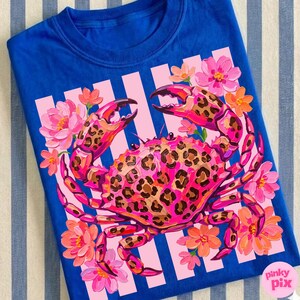 May include: A royal blue t-shirt featuring a vibrant crab design. The crab is pink with a leopard print pattern, surrounded by pink and orange flowers. The design is set against a pink and white striped background.