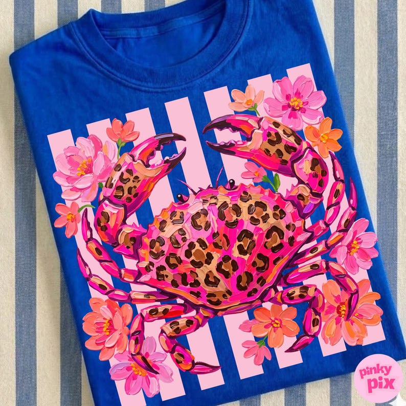 May include: A royal blue t-shirt featuring a vibrant crab design. The crab is pink with a leopard print pattern, surrounded by pink and orange flowers. The design is set against a pink and white striped background.
