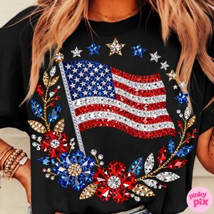 May include: Black t-shirt featuring a sparkling American flag design with red and white stripes and blue stars. The flag is surrounded by a wreath of red, white, and blue floral and star embellishments. A patriotic themed apparel item.