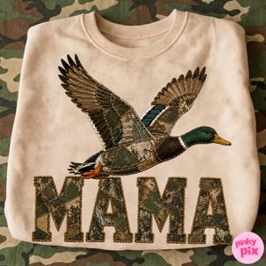 May include: A light beige crewneck sweatshirt featuring a large embroidered mallard duck in flight above the word MAMA. The duck and the letters are filled with a patchwork camouflage pattern in shades of green, brown, and tan.