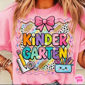 May include: Pink t-shirt with a colorful "KINDERGARTEN" graphic. The design includes a pink bow, a pencil, a notepad, and a pencil case with school supplies. The graphic is framed with a decorative border.