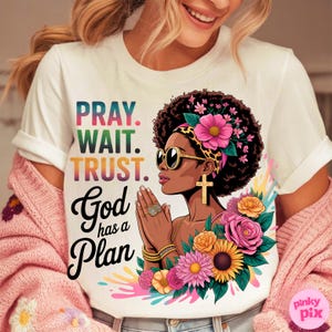 May include: White t-shirt with the text "PRAY. WAIT. TRUST. God has a Plan" and a graphic of a woman with sunglasses, a cross earring, and floral accents. The design features vibrant colors and a spiritual theme.