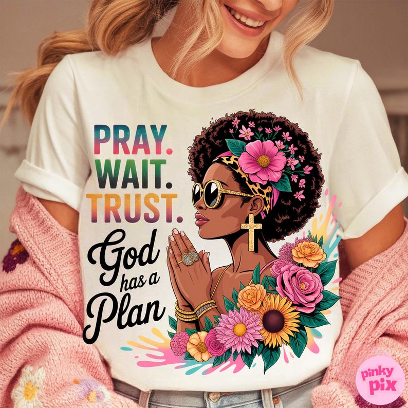 May include: White t-shirt with the text "PRAY. WAIT. TRUST. God has a Plan" and a graphic of a woman with sunglasses, a cross earring, and floral accents. The design features vibrant colors and a spiritual theme.