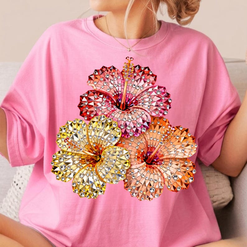 May include: Pink t-shirt featuring a vibrant floral design. The graphic showcases three hibiscus flowers in shades of red, yellow, and orange, adorned with sparkling details. The shirt has short sleeves and a crew neck.