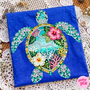 May include: A blue t-shirt featuring a bejeweled sea turtle design. The turtle has a turquoise shell and limbs, with a yellow and gold border. The turtle's shell has a tropical scene with pink and white flowers, coral, and ocean waves.