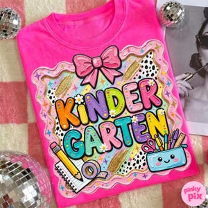 May include: Bright pink t-shirt with a colorful "Kindergarten" graphic. The design includes a pink bow, gold accents, and school supplies like a pencil, notebook, and a pencil case. The text is in multiple colors with flower accents.