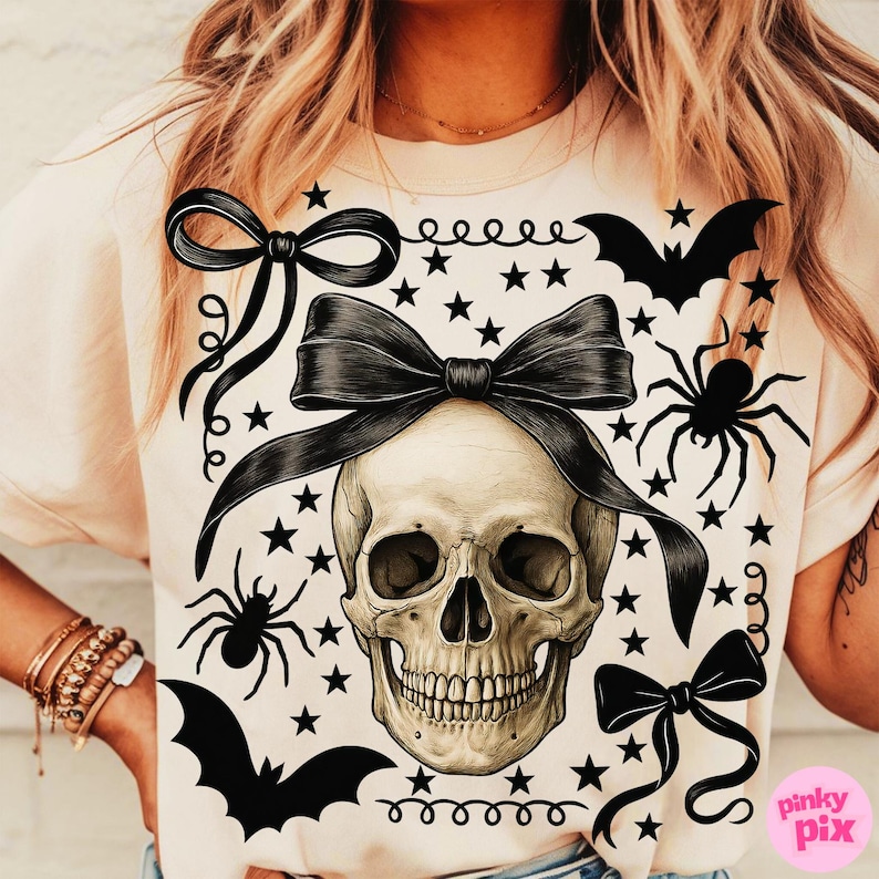 May include: A light beige t-shirt featuring a skull with a large black bow, surrounded by black bows, bats, spiders, and stars. The design has a Halloween theme.