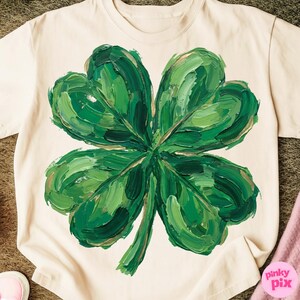 K&ouml;nnte beinhalten: Cremefarbenes T-Shirt mit einem gro&szlig;en, strukturierten vierbl&auml;ttrigen Kleeblatt-Design. Das Kleeblatt ist in Gr&uuml;nt&ouml;nen mit goldenen Akzenten bemalt, was einen k&uuml;hnen und k&uuml;nstlerischen Look erzeugt. Das Shirt ist ein Freizeitkleidungsst&uuml;ck.
