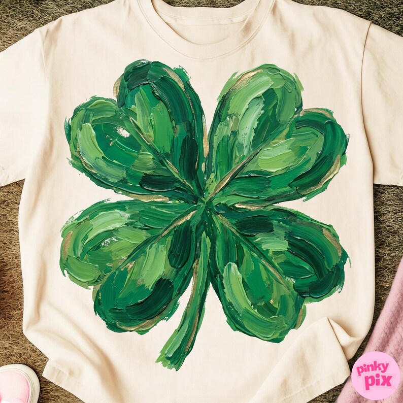 K&ouml;nnte beinhalten: Cremefarbenes T-Shirt mit einem gro&szlig;en, strukturierten vierbl&auml;ttrigen Kleeblatt-Design. Das Kleeblatt ist in Gr&uuml;nt&ouml;nen mit goldenen Akzenten bemalt, was einen k&uuml;hnen und k&uuml;nstlerischen Look erzeugt. Das Shirt ist ein Freizeitkleidungsst&uuml;ck.