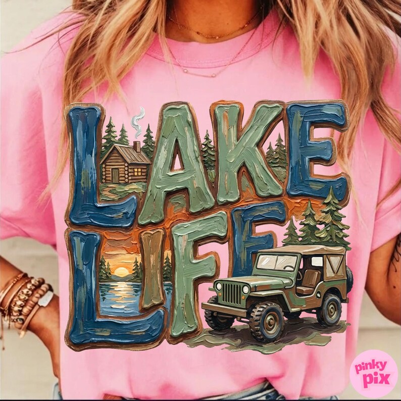 May include: Pink t-shirt with the words "LAKE LIFE" in a 3D design. The design includes a cabin, trees, a lake with a sunset, and a green jeep. The shirt has short sleeves and a round neckline.