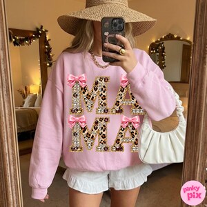May include: Pale pink sweatshirt featuring the word "MAMA" in a leopard print pattern, adorned with rhinestones and pink bows. This item is part of a clothing range.
