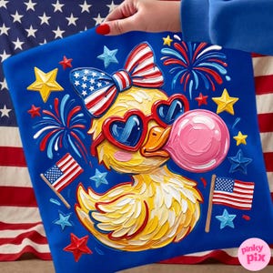 May include: A blue graphic tee featuring a yellow duck wearing heart-shaped sunglasses and a bow tie, both in red, white, and blue. The duck is blowing a pink bubble, surrounded by stars, fireworks, and American flags. The design has a textured, painted look.