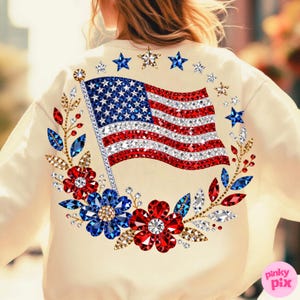 May include: Cream-colored sweatshirt featuring a sparkling American flag design. The flag is embellished with red, white, and blue rhinestones. A floral wreath with red, blue, and clear gems surrounds the flag, with star accents.