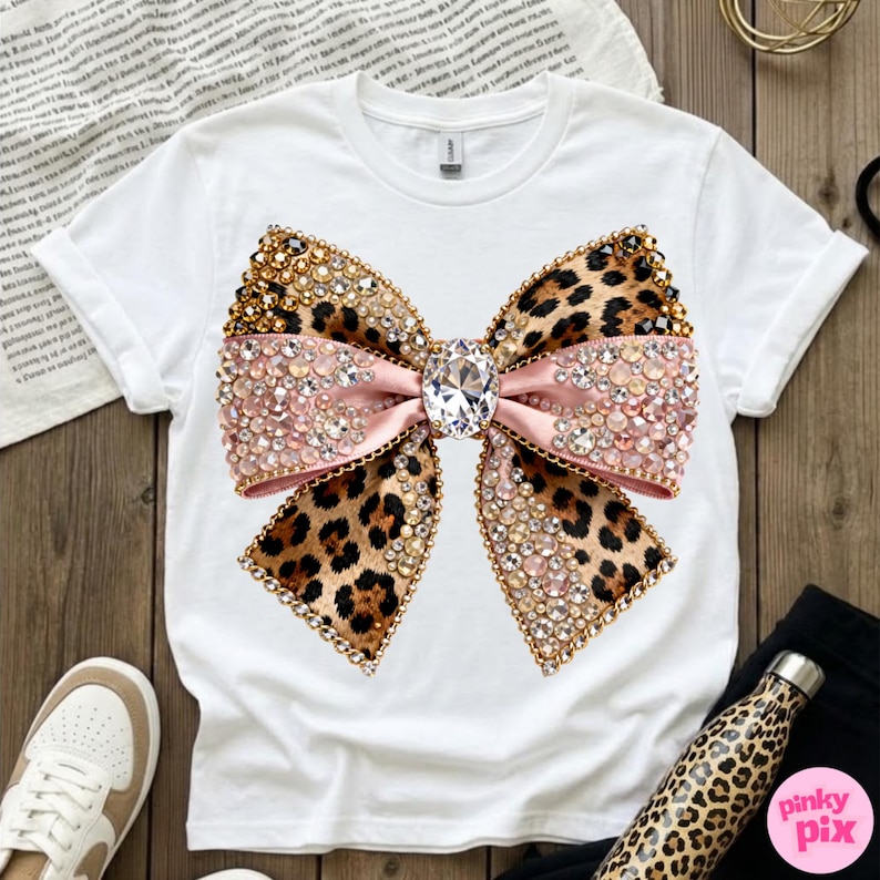 May include: White t-shirt featuring a large bow design. The bow has a leopard print pattern with pink ribbon, and is embellished with rhinestones and gold accents. A large, clear gem is in the center of the bow.