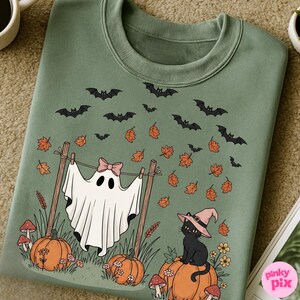 May include: Sage green sweatshirt featuring a Halloween-themed design. The graphic includes a ghost with a pink bow, pumpkins, a black cat wearing a witch hat, bats, and autumn leaves. The design is whimsical and festive.