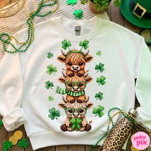 May include: White sweatshirt featuring a stack of three cartoon Highland cows, each adorned with shamrocks and glasses. The cows are brown with green accents, surrounded by green shamrock graphics. A festive St. Patrick's Day theme.