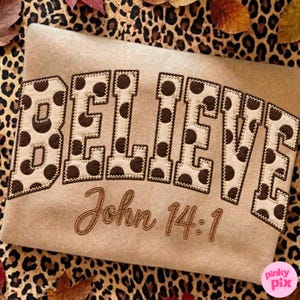 Könnte beinhalten: Ein beigefarbenes Sweatshirt mit dem Wort BELIEVE in großen Blockbuchstaben mit einem cremefarbenen und braunen Punktemuster. Unter dem Wort ist der Text John 14:1 in brauner Schreibschrift gestickt. Das Kleidungsstück hat einen sichtbaren Saum.